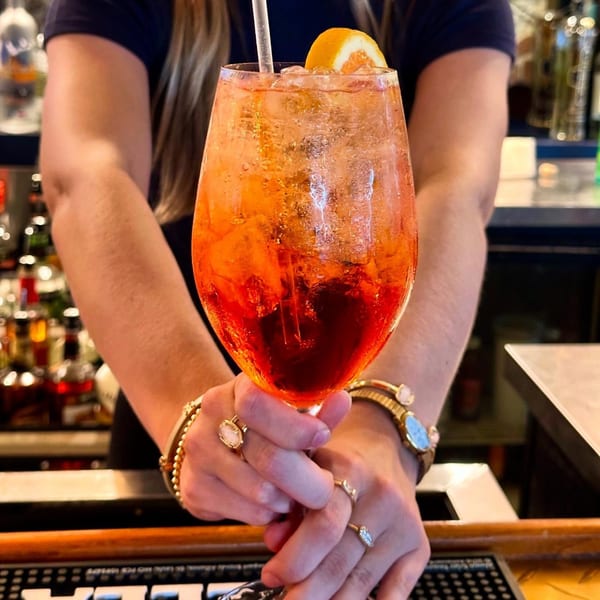 Bartender's hands holding a large glass of orange cocktail with ice and an orange slice.