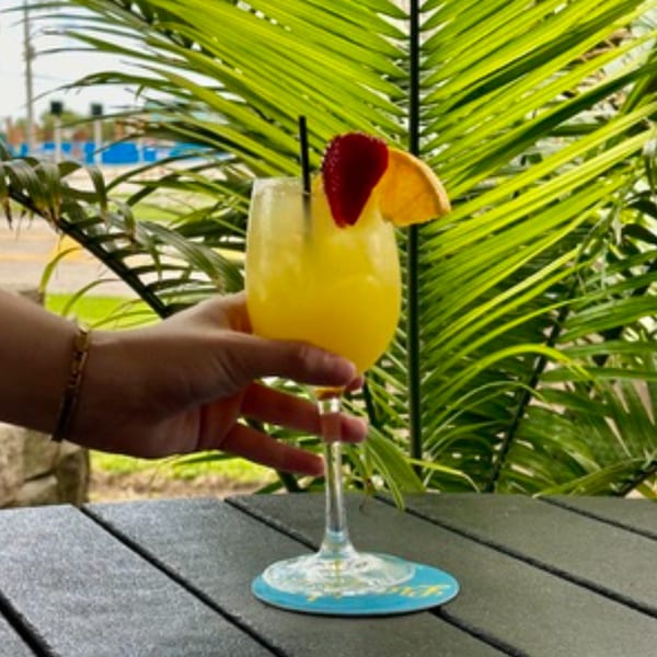 Hand holding a yellow cocktail garnished with strawberry and orange, against a palm leaf background.