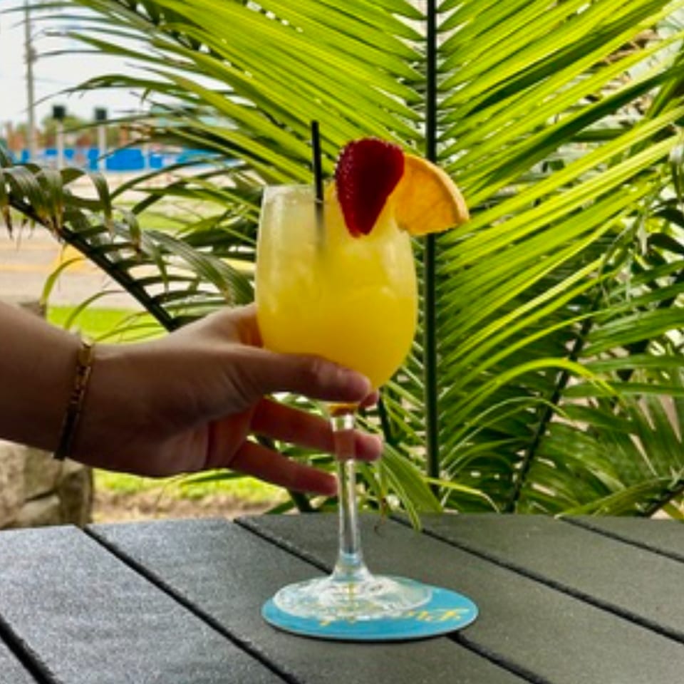 Hand holding a yellow cocktail garnished with strawberry and orange, against a palm leaf background.