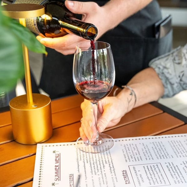 A server pours red wine into a glass for a customer at a table with a menu.