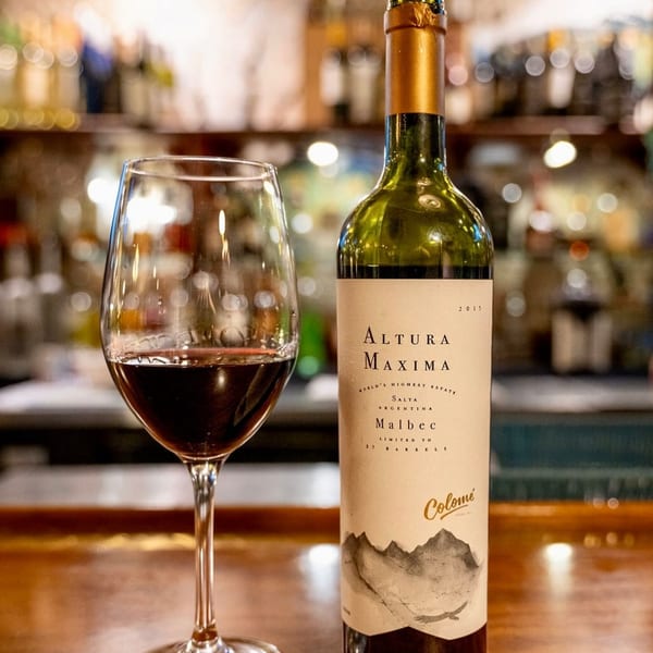A glass of red wine next to a bottle of Altura Maxima Malbec on a wooden bar.