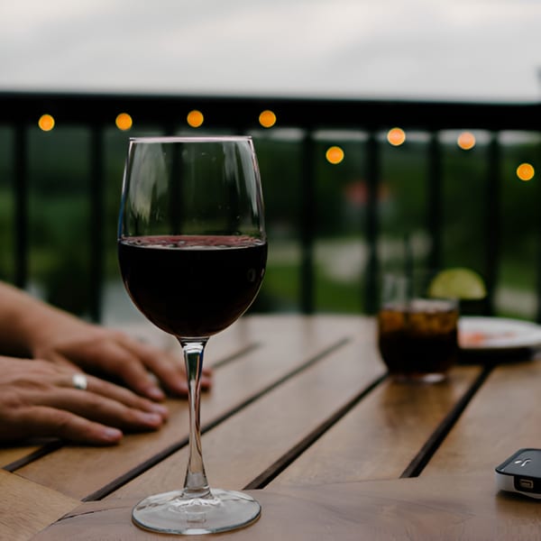 A glass of red wine on a wooden table with string lights and a person's hands in the background.
